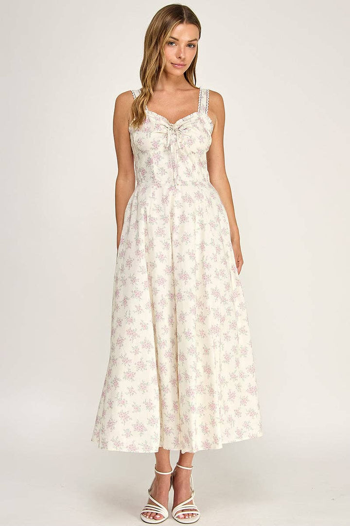 A woman modeling a sleeveless white maxi dress with a delicate pink floral pattern, featuring a sweetheart neckline with a bow detail and a flared skirt, set against a plain light gray studio background.