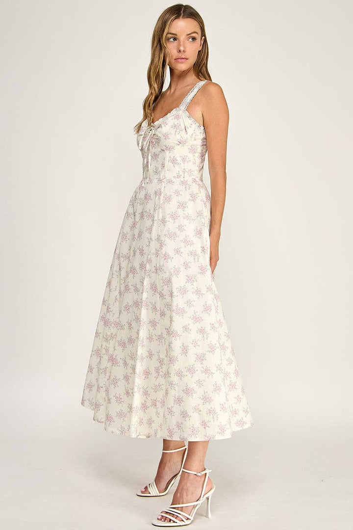 A woman modeling a sleeveless midi dress with a white base and a light pink floral pattern, paired with white strappy high-heeled sandals against a plain light gray studio background.