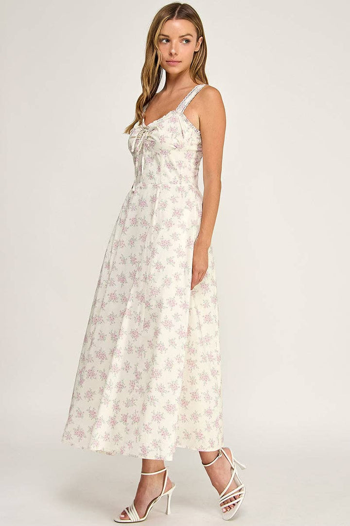 A woman modeling a sleeveless midi dress with a white base and a light pink floral pattern, featuring a tie-neck detail and thin shoulder straps, paired with white strappy high-heeled sandals against a light gray studio background.
