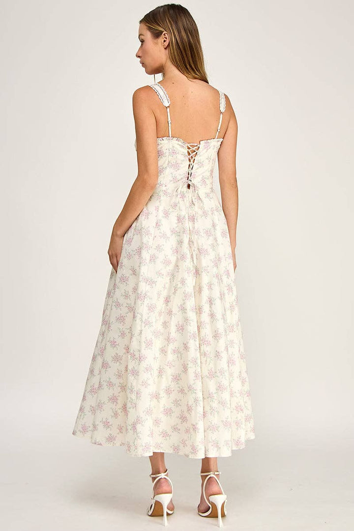 A woman modeling a sleeveless maxi dress from the back, featuring a cream base with a light pink floral pattern and lace-up detailing at the back, set against a plain light gray studio background.
