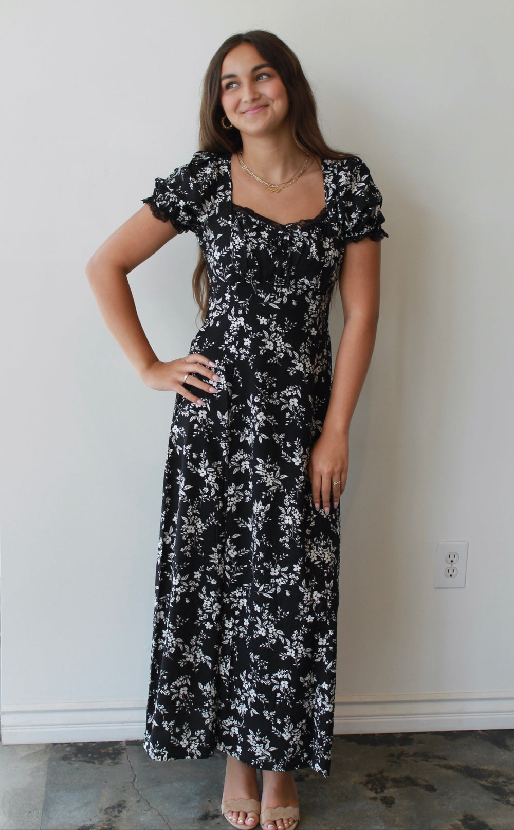 Woman wearing a black floral dress standing against a white wall.