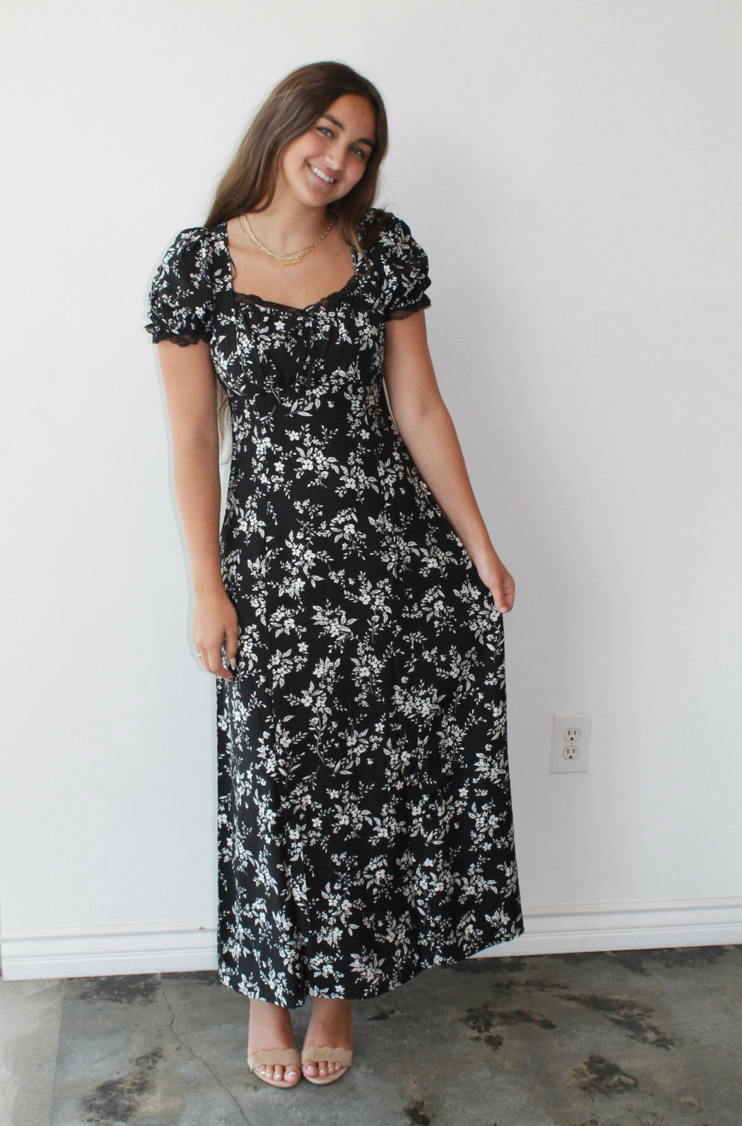 Woman wearing a black floral dress standing against a white wall.