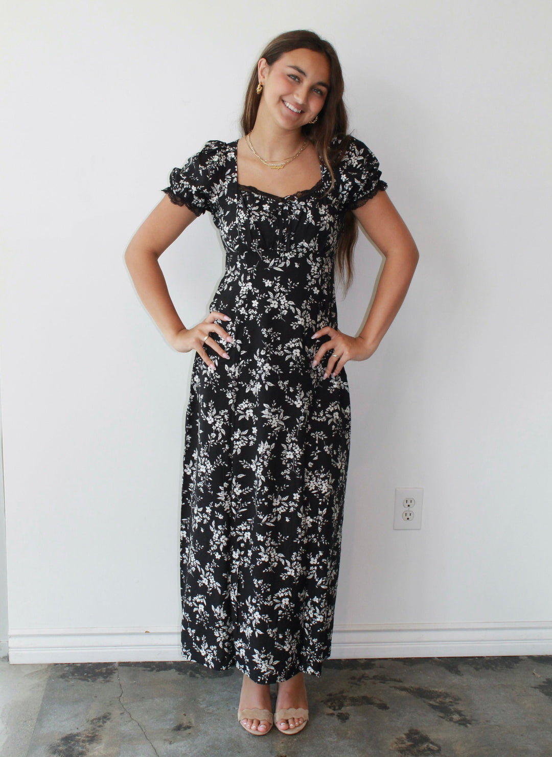 Woman wearing a black floral dress standing against a white wall.