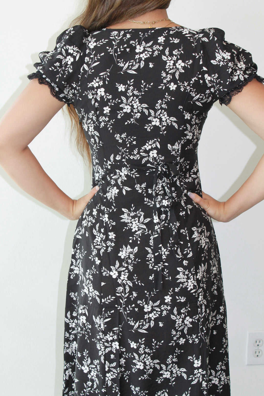 Black floral dress worn by a person with hands on hips against a white wall.
