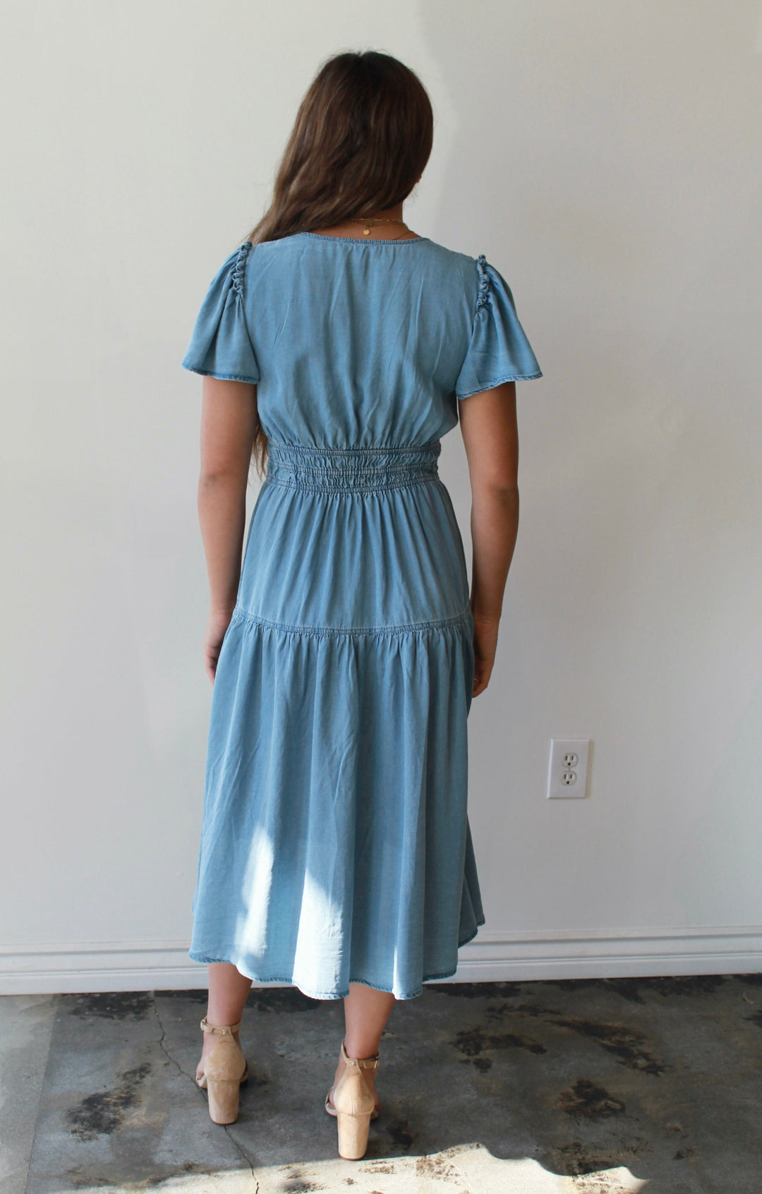 Woman wearing a blue dress standing in a room with a white wall and window.